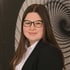 Junge Frau mit langen braunen Haaren, Brille, weißem Hemd und schwarzem Blazer lächelt vor spiralförmigem Hintergrund.