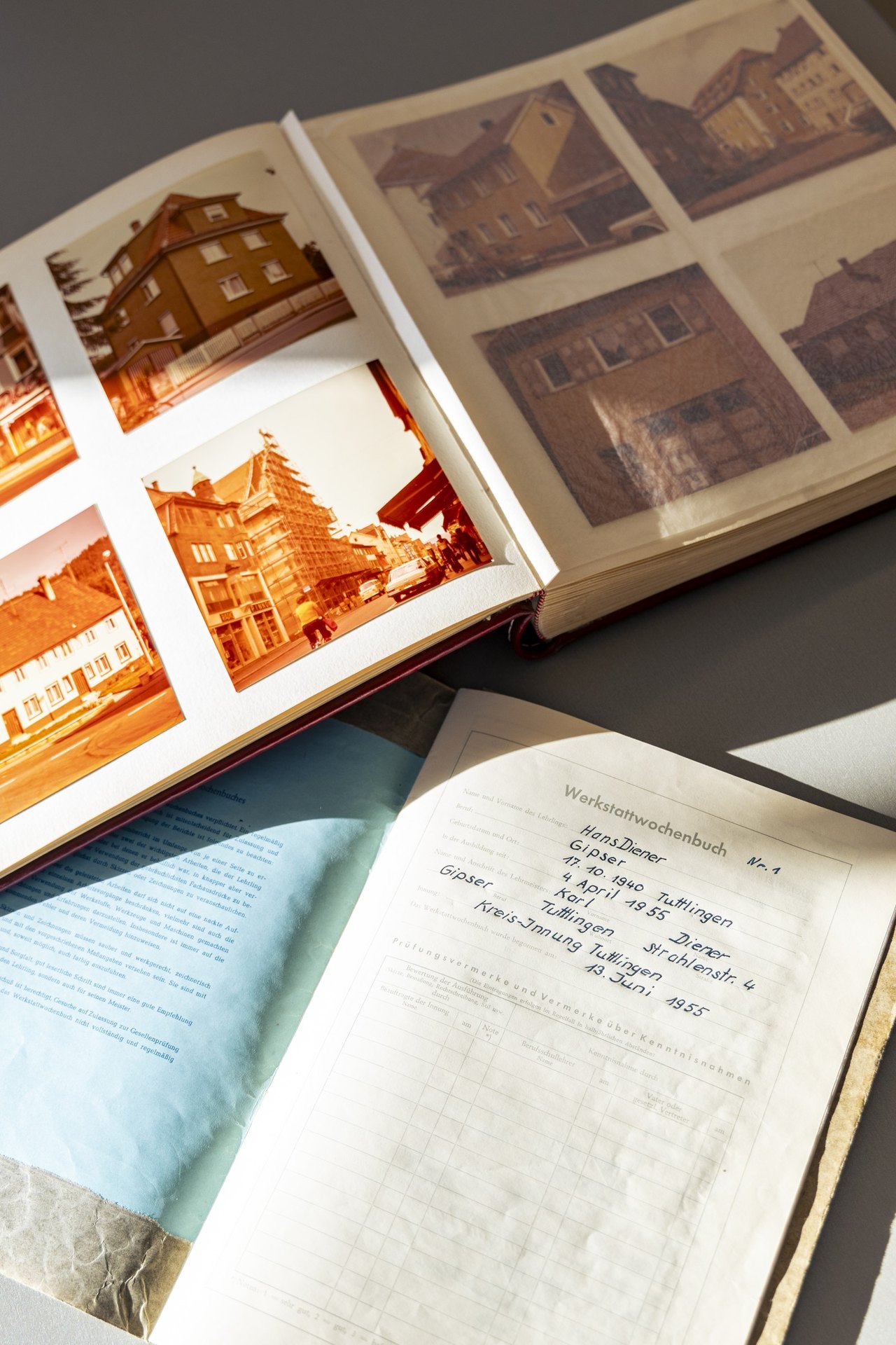 Aufgeschlagenes Fotoalbum mit alten Gebäudeaufnahmen, davor das Werkstattwochenbuch des Lehrlings Hans Diener von 1955