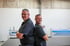 Andreas und Norman Singer in schwarzen Poloshirts stehen Rücken an Rücken in einer Werkstatt vor einer Metallbiegemaschine.