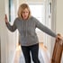 A woman in a gray hoodie leans against a wall and holds a wooden chair for support in a hallway.