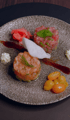 Zwei Tartar-Anrichten, eine aus Lachs und eine aus rotem Fleisch, auf einem grauen Teller mit Soße, Tomaten und Ingwer.