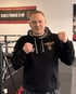 Mann in schwarzem Hoodie mit „FIGHTCLUB LANGENAU“ Logo, Fäuste erhoben, vor einem Boxring.