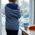 Woman in a grey cardigan hugging herself by a window next to a steaming mug and folded orange blanket.