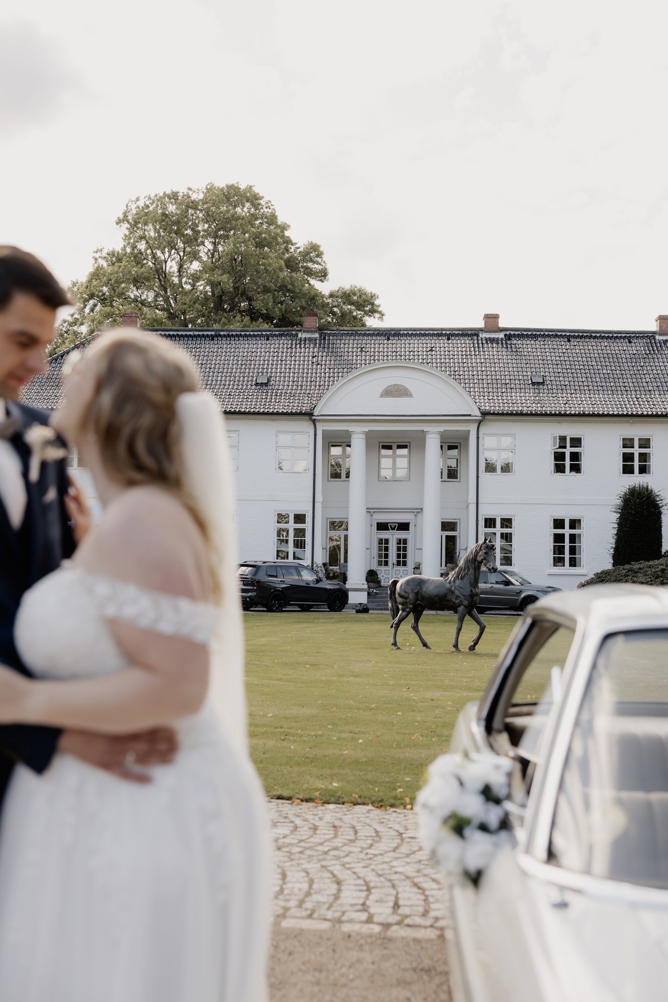 Bräutigam hält die Braut im Arm, im Hintergrund Gutshaus und Oldtimer