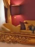 Detailansicht eines barocken Goldspiegels, der ein Zimmer mit roter Wand, Stehlampe und beigem Sofa reflektiert.