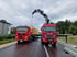 Zwei rote Lastwagen mit roten Logos und Containern stehen auf einer asphaltierten Fläche unter bewölktem Himmel.