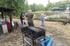 Personen stehen oder sitzen auf einem Bahnhofsgelände mit Grill, Schienen und einem Auto im Hintergrund.