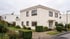 Zweistöckiges Einfamilienhaus mit Flachdach, heller Fassade, braunen Fenstern und gepflegtem Vorgarten an einer Straße.