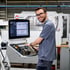 Ein lächelnder Techniker mit Brille bedient ein CNC-Maschinen-Bedienfeld mit Bildschirm