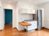 Moderne kleine Küchenzeile mit weißen Schränken, Edelstahlkühlschrank, Holztisch und Blick in einen Raum mit blauer Wand.