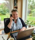 Lächelnde Frau mit kurzen Haaren telefoniert am Schreibtisch mit Laptop vor einem Fenster mit Gartenblick.
