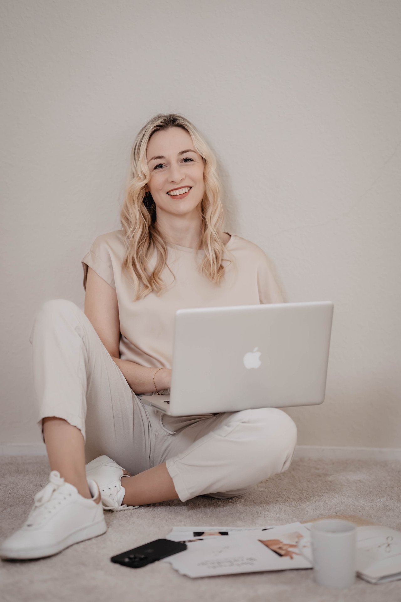 Lächelnde blonde Frau sitzt auf dem Boden und arbeitet mit einem Laptop auf dem Schoß.