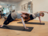 Zwei Frauen führen die Pilates-Übung Seitstütz auf schwarzen Matten in einem hellen Studio mit Trainingsgeräten aus.