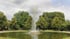 Hohe Wasserfontäne in einem runden Teich, umgeben von grünen Bäumen unter bewölktem Himmel.