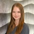 Junge Frau mit langen rotbraunen Haaren lächelt vor einer grauen Wand mit Wellenmuster.
