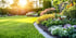 Gepflegter Garten mit grünem Rasen, Blumenbeet und strahlender Sonne am Horizont.