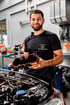KFZ-Mechaniker vom Autohaus Wetterauer hält ein schwarzes Motorbauteil mit roter Dichtung über einem geöffneten Motorraum.