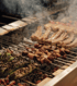 Meat skewers, peppers, and other food grilling over hot coals with smoke rising.