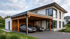 Modernes zweistöckiges Wohnhaus mit weißer Fassade und angebautem Holz-Carport, in dem zwei Autos parken.
