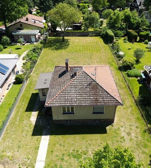 Luftaufnahme eines Hauses mit Ziegeldach und großem Rasengrundstück, umgeben von anderen Häusern und Bäumen.