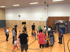 Menschen und Kinder spielen in einer Turnhalle mit Holzwänden und orangefarbenem Boden.