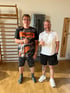 Zwei lächelnde Männer zeigen den Daumen nach oben in einem Fitnessraum mit Sprossenwand und Trainingsgeräten.