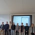 Sieben Studenten und ein Professor stehen vor einer großen Leinwand, die eine Webseite anzeigt.