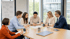 Sechs Geschäftsleute sitzen um einen Konferenztisch, betrachten Dokumente und diskutieren.