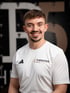 Lächelnder Physiotherapeut im weißen Sportshirt mit „THERAPIER this is PHYSIO“-Logo vor dunklem Hintergrund und Pier5.