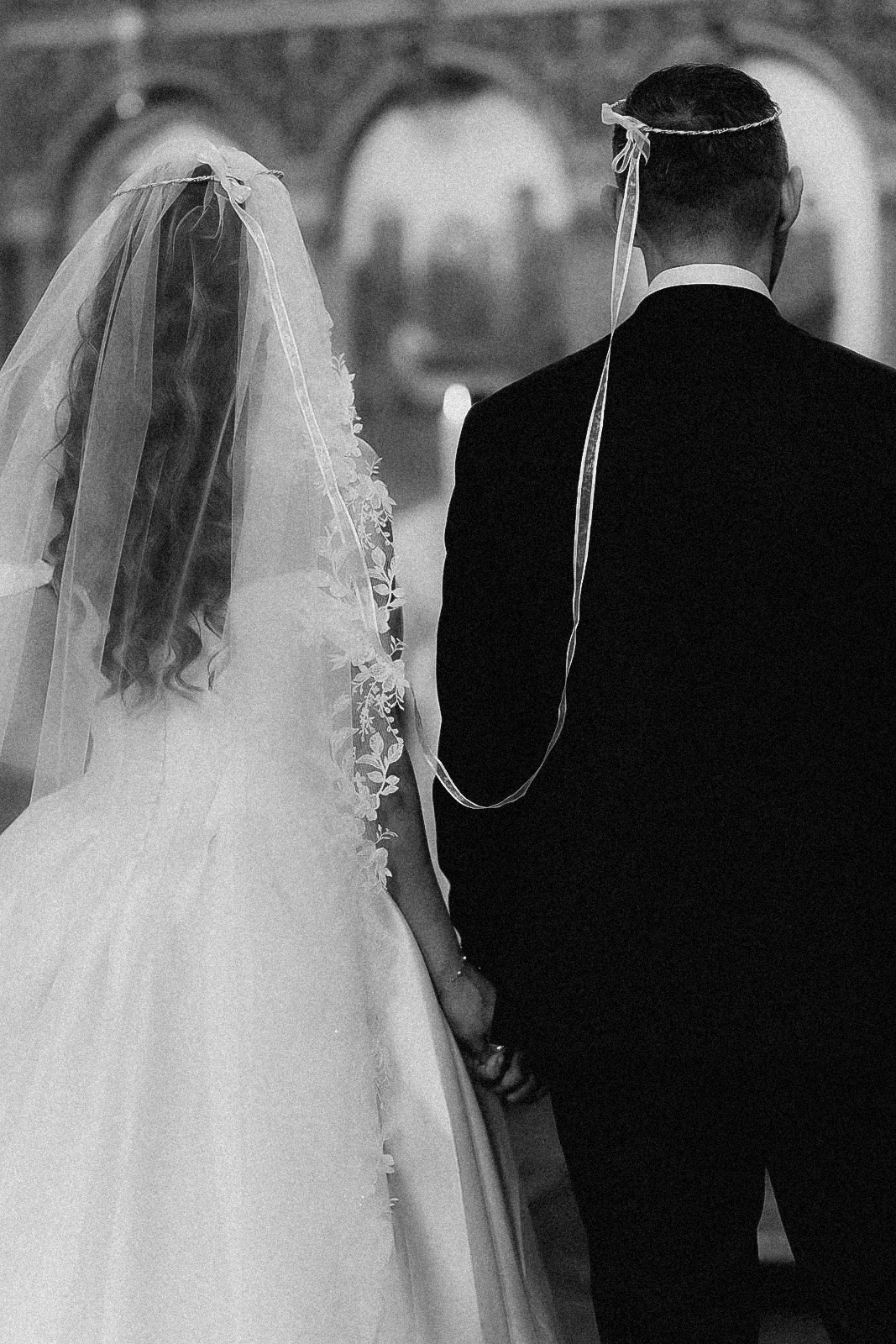 Schwarz-Weiß-Aufnahme: Das Brautpaar steht von hinten am Altar, beide tragen die mit einem Band verbundenen Ehekronen, während der Trauung in einer orthodoxen Kirche in Köln.