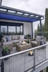 Moderne Terrasse mit grauen Outdoor-Möbeln, blauer Pergola, hellem Fliesenboden und Glasgeländer.