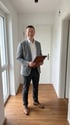 Lächelnder Mann im grauen Sakko steht mit Klemmbrett in einem hellen Flur mit Laminatboden.