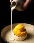 Eine Hand gießt helle Soße auf einen Reiskuchen mit gelber Mango-Glasur und schwarzen Samen.
