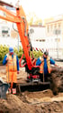 Zwei Bauarbeiter in Warnwesten arbeiten mit einem orangefarbenen Kubota-Bagger an einem tiefen Erdaushub auf einer Baustelle.