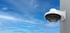 Weiße Hanwha Dome-Überwachungskamera an einer grauen Außenwand vor blauem Himmel mit Wolken.