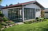 Kleines, weißes Gartenhaus mit Falttüren aus Glas und roten Ziegeln, umgeben von einem grünen Garten mit Blumen.