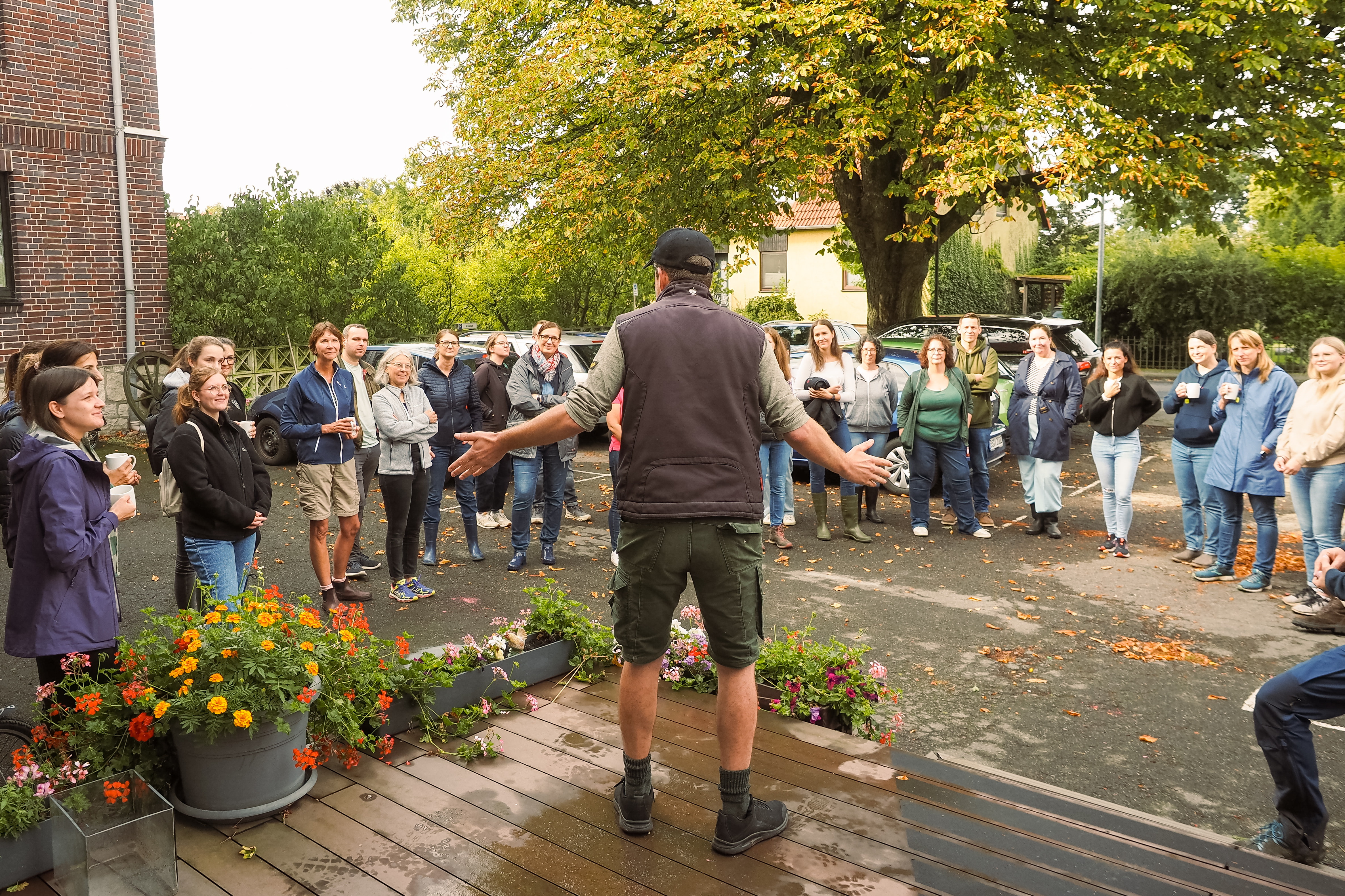 Mann mit Baseballkappe spricht vor einer Gruppe von Frauen auf einem Parkplatz mit Bäumen und Blumen.