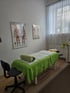 Behandlungsraum mit grüner Massageliege, anatomischen Postern an der Wand und einem Rollhocker auf Laminatboden.