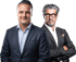 Zwei Männer in Anzügen, der linke lächelt, der rechte mit Brille und Bart blickt ernst in die Kamera.