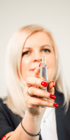 Blonde Frau mit roten Nägeln hält ein silbernes Permanent-Make-up-Gerät in die Kamera.