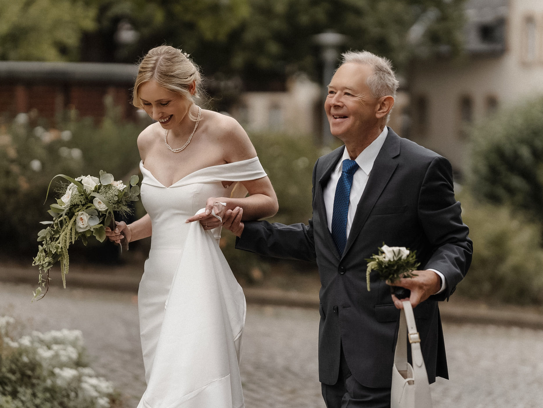 Braut geht lachend Arm in Arm mit ihrem Vater zur Trauung am Schloss Eller in Düsseldorf.