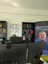 Büro mit zwei schwarzen Regalen, Schreibtischstuhl, zwei Monitoren und einem Roll-up-Display.