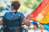 Person im Rollstuhl mit Regenbogenfahne auf der Schulter, von hinten gesehen.