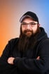 Mann mit langem Bart, schwarzer Kappe und Brille, der in einem schwarzen Hoodie die Arme verschränkt.