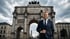 Andreas Hatzl im Anzug lächelt vor dem Siegestor in München unter einem bewölkten Himmel.