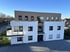 Modernes Mehrfamilienhaus mit weißer und brauner Fassade, Balkon und dunklen Fenstern unter blauem Himmel.