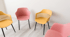 Vier leere Stühle in Rosa und Gelb, im Kreis angeordnet, vor einer weißen Wand.