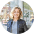 Lächelnde Frau mit blonden Locken und schwarzer Lederjacke vor verschwommenem Stadthintergrund.
