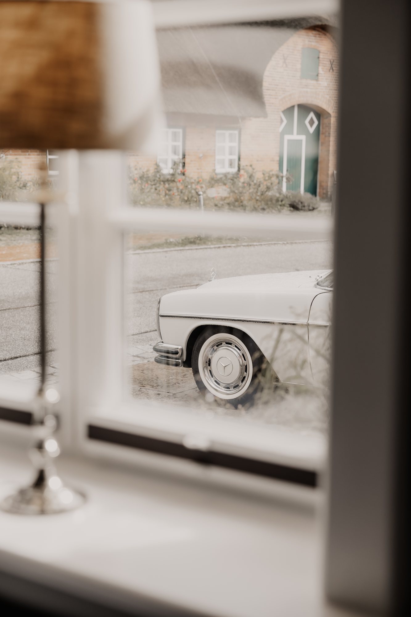 Blick durch ein Fenster auf einen weißen Oldtimer vor einem Gutshaus