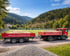 Roter LKW zieht Anhänger mit Sand auf Schotterweg vor bewaldeten Bergen und Fußballfeld.